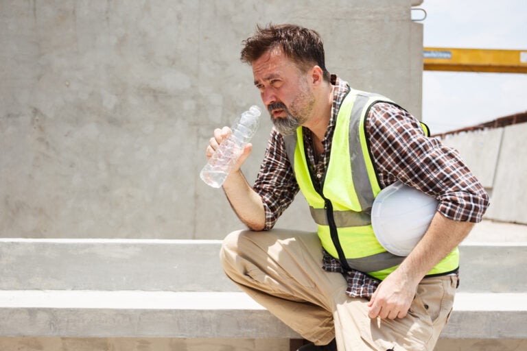 Dangerous from heat wave during working outside the building. Caucasian male construction worker resting and drinking water from bottle in a very hot day. Heat stroke health problem.